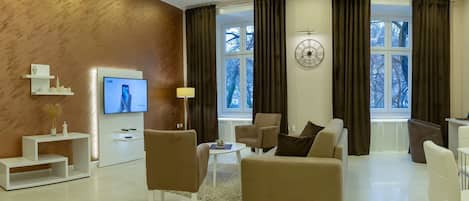 A modern living room with a flat-screen TV, a clock, and a view of trees through large windows.