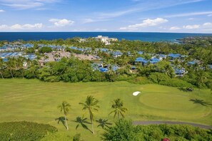 A golf course with a large building in the background, surrounded by palm trees and blue-roofed structures. - Fully Renovated Luxury Condo | Ocean Views + AC (Kailua-Kona)