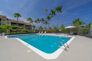 A swimming pool with a clear blue water, surrounded by palm trees and a building with a tiled roof. - Fully Renovated Luxury Condo | Ocean Views + AC (Kailua-Kona)