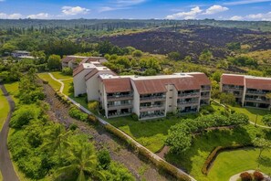 A modern hotel with multiple floors, a central courtyard, and a lush green landscape with hills in the background. - Fully Renovated Luxury Condo | Ocean Views + AC (Kailua-Kona)