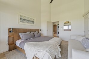 A bedroom with a bed, bedside tables, a lamp, a large window, and a view of a laundry area. - Fully Renovated Luxury Condo | Ocean Views + AC (Kailua-Kona)