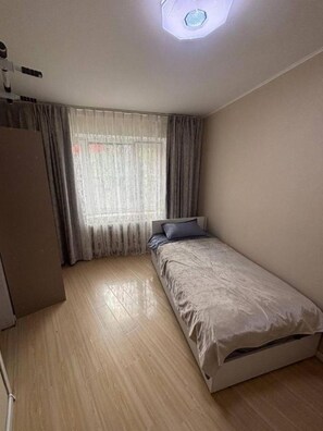 Ein kleines Zimmer mit einem Einzelbett, Holzboden, einem Bett mit grauer Matratze und blauem Kopfkissen, einem Fenster mit grauen Vorhängen und einer Deckenleuchte.