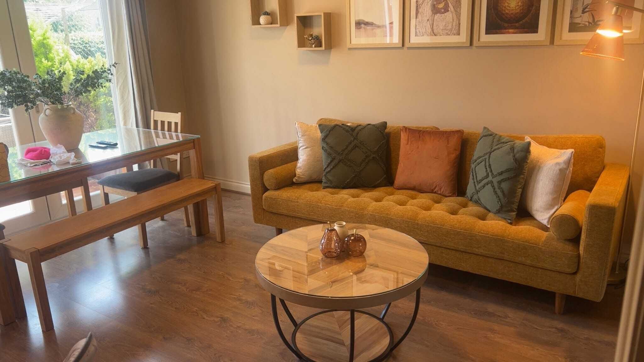 Una sala de estar con un sofá, una mesa de centro, una mesa de comedor con sillas y una ventana con plantas.