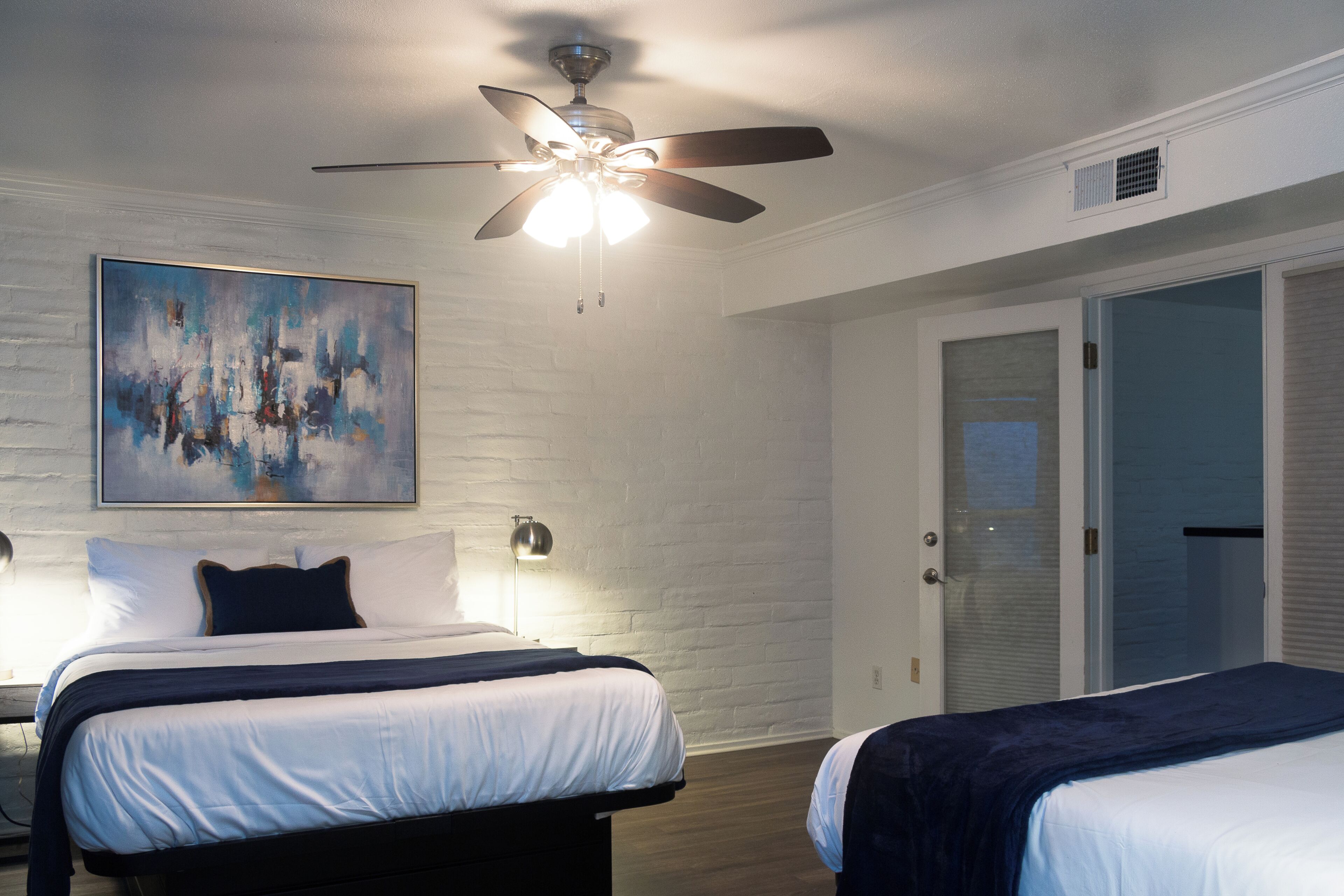 A hotel room with a bed, a ceiling fan, a painting on the wall, and a door.