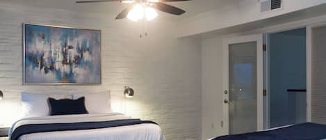 A hotel room with a bed, a ceiling fan, a painting on the wall, and a door.