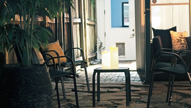 Uma área externa com mesas e cadeiras, um vaso de planta e uma porta que dá para um espaço interno.