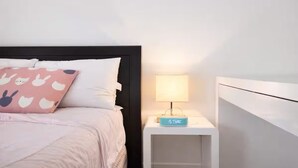 En pænt redt seng med et hvidt sengegærde, en lyserød pude med kaninmønster og et natbord med en lampe og et ur.