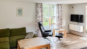 Ett vardagsrum med en grön soffa, ett soffbord i trä, en svart läderfåtölj, ett litet sidobord i trä och en tv som står på ett vitt skåp.