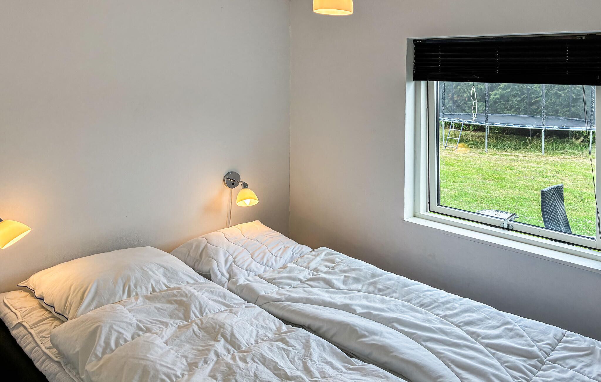Un lit simple avec du linge de lit blanc, une lampe fixée au mur et une fenêtre donnant sur un espace verdoyant.