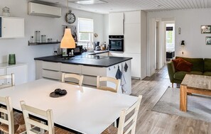 A modern kitchen with a central island, a dining area with a white table and chairs, and a living room with a green sofa and a wooden coffee table. - 4 bedroom awesome home in Væggerløse (Væggerløse)