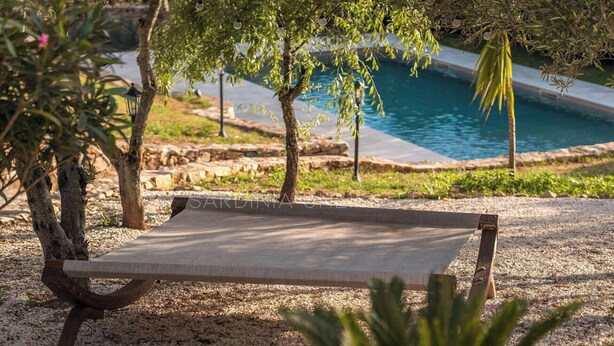 A wooden bench with a metal frame, positioned on a gravel surface near a poolside area with trees and a clear view of the water. - Villa Le Sfumature Mediterranee - Sardinia Unlimited (San Teodoro)