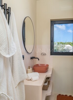 A bathroom with a large oval mirror, a brown sink, and a window with a view of greenery. - Villa Le Sfumature Mediterranee - Sardinia Unlimited (San Teodoro)