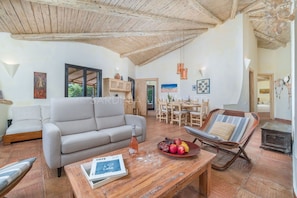 Un vaste espace de vie comprenant un canapé, une table à manger et un fauteuil. Le plafond est en bois et la pièce offre une vue sur l’extérieur.