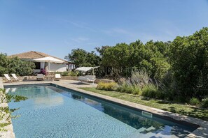 Ein Swimmingpool mit Liegestühlen und Sonnenschirmen, umgeben von viel Grün und einem Steinhaus.