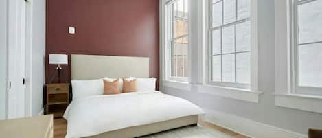 A bedroom with a large bed, a nightstand, a lamp, and a window with a view of the outside.