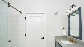 Ein modernes Badezimmer mit einer weißen Tür, einer Duschkabine aus Glas, einem Waschbecken mit Goldarmatur und einem Spiegel mit goldener Rahmung.