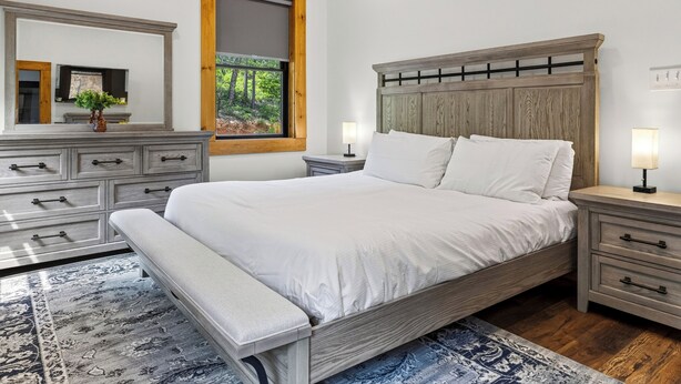 Un dormitorio con cama, mesitas de noche, un banco, cómoda y un espejo. Se ve un ventanal con vista a árboles.