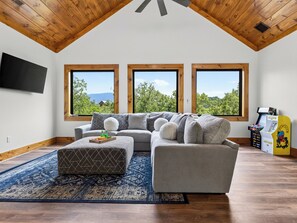 A modern living room with a sectional sofa, ottoman, and arcade games, featuring large windows and a wooden ceiling. - Sleeps 20 + Indoor Pool + Game Room Near Pigeon Forge | Spurling Retreat (Sevierville)