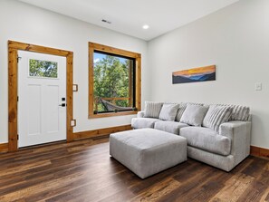 Una habitación con puerta de madera, un ventanal con vista a árboles, un sofá gris y un cuadro en la pared.