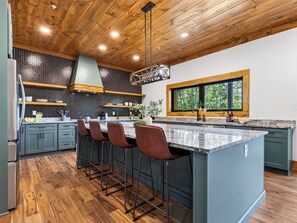 A modern kitchen with a central island, bar stools, wooden ceiling, and a window with a view of trees. - Sleeps 20 + Indoor Pool + Game Room Near Pigeon Forge | Spurling Retreat (Sevierville)