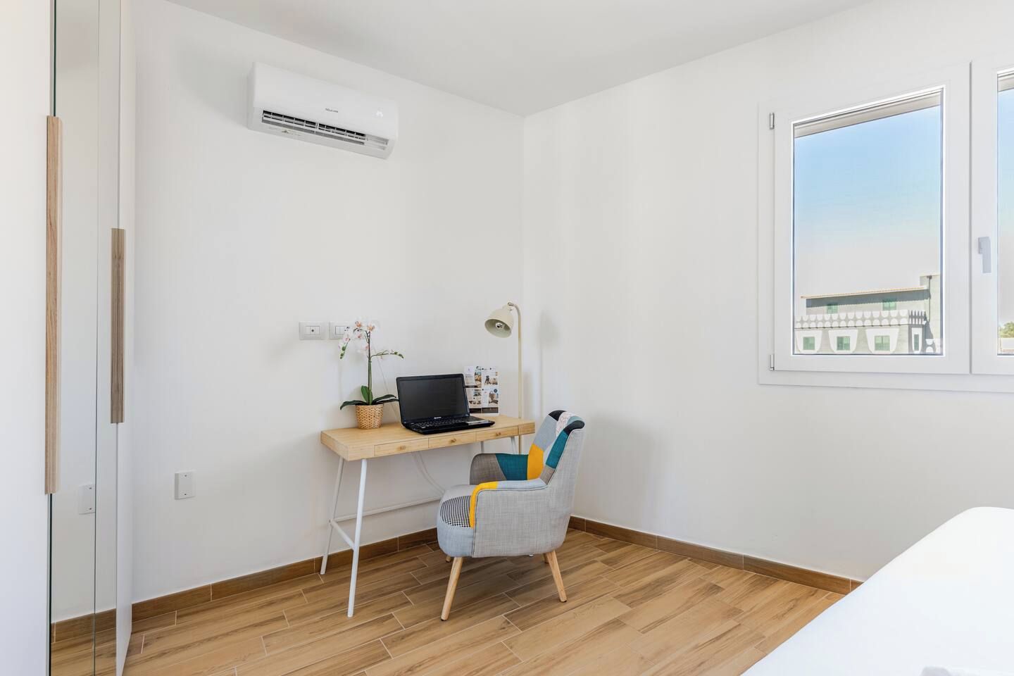 A room with a wooden desk, a laptop, a chair, an air conditioner, and a window with a view.