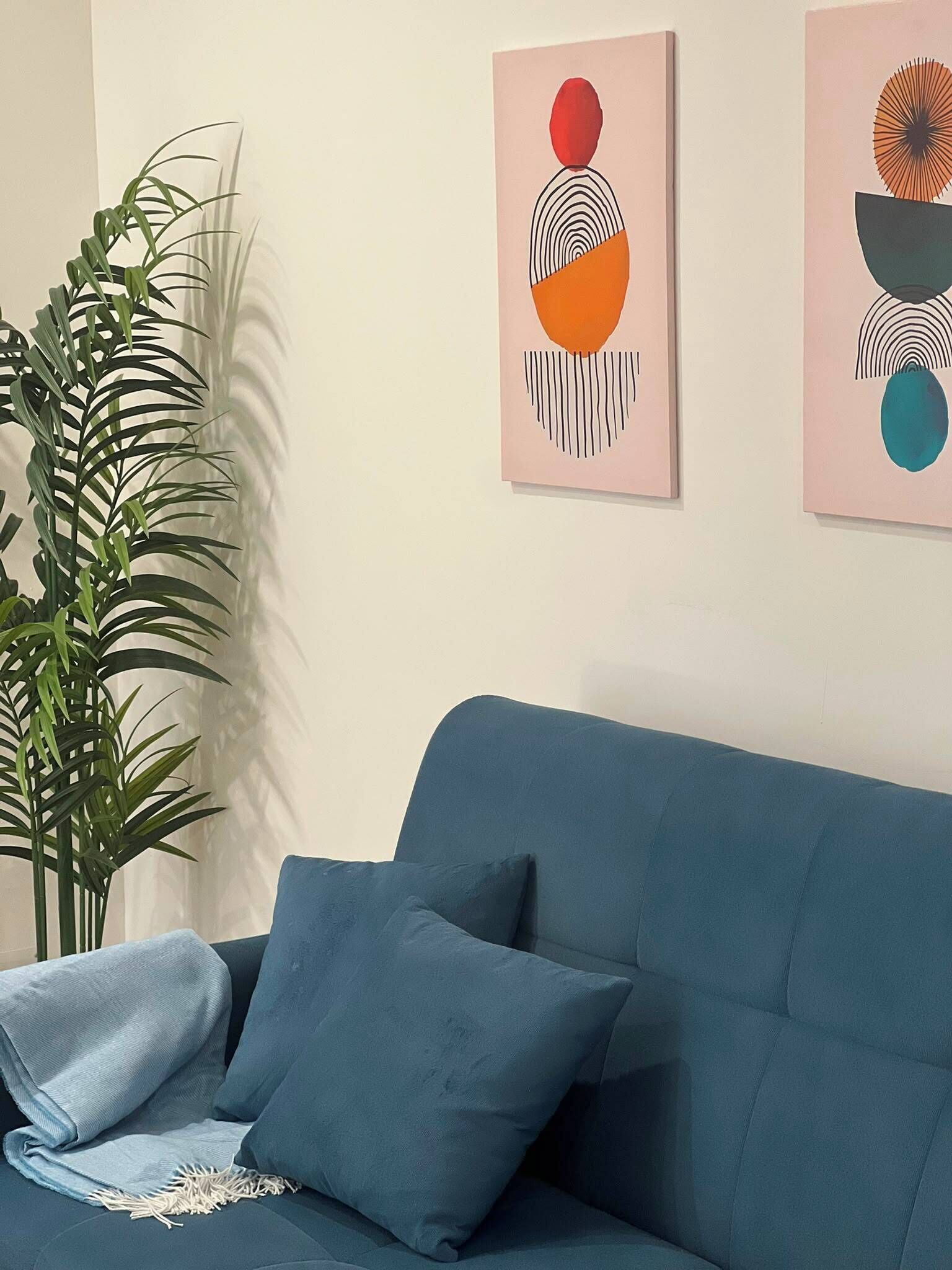 Ein blaues Sofa mit zwei Kissen, einer Topfpflanze und abstrakter Kunst an der Wand.
