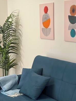 A blue sofa with two cushions, a potted plant, and abstract art on the wall.