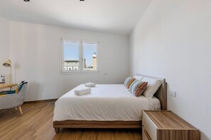 Quarto com cama, escrivaninha, cadeira, janela e vista para prédios.
