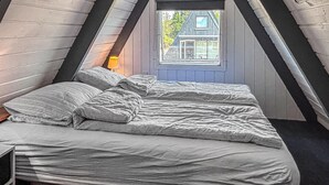 Et soveværelse med skråvæg, en seng med hvid dyne og puder, en sengetavlelampe og et vindue med udsigt til et hus.