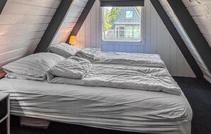 A bedroom with a sloped ceiling, a bed with a white duvet and pillows, a bedside lamp, and a window with a view of a house. - Gorgeous home in Farsø with WiFi (Farsø)