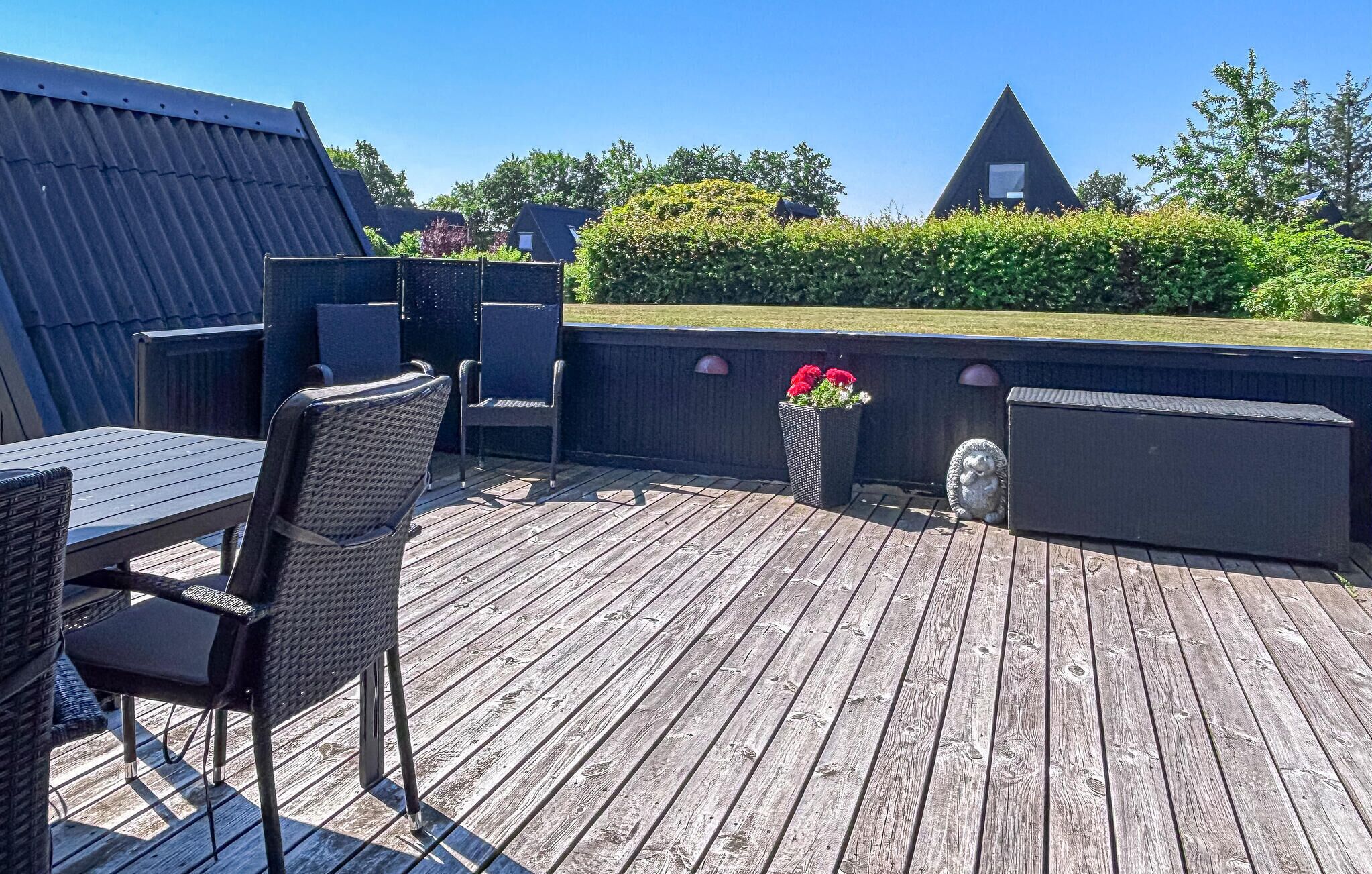 Eine Holzterrasse mit Gartenmöbeln, einem Blumenbeet und einem dreieckigen Gebäude im Hintergrund.