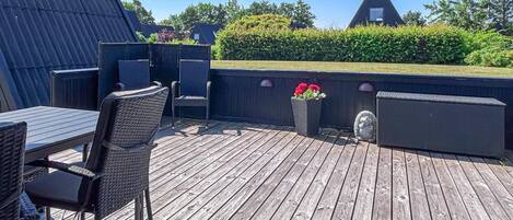Eine Holzterrasse mit Gartenmöbeln, einem Blumenbeet und einem dreieckigen Gebäude im Hintergrund.