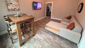 En matplats med ett träbord och stolar, en soffa, en tv som är monterad på väggen och en hall som leder till ett annat rum.