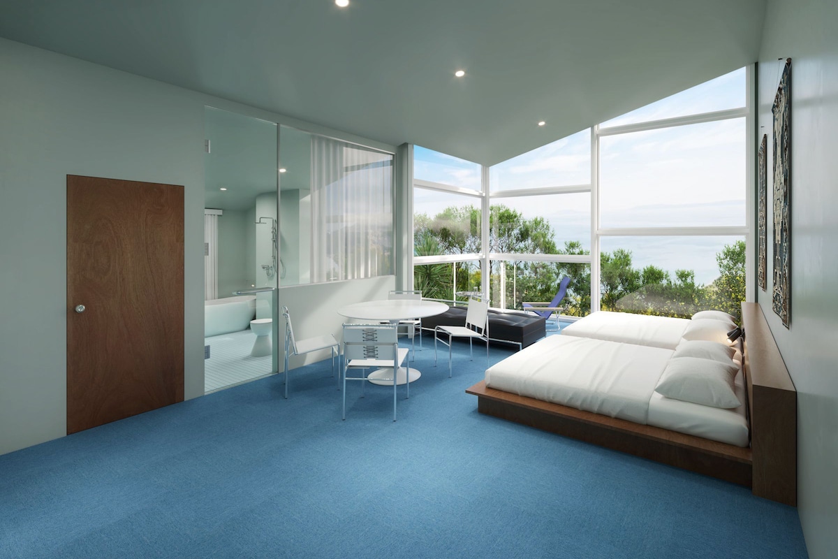 A modern bedroom with a large bed, a desk, and a chair, featuring a large window with a view of greenery.
