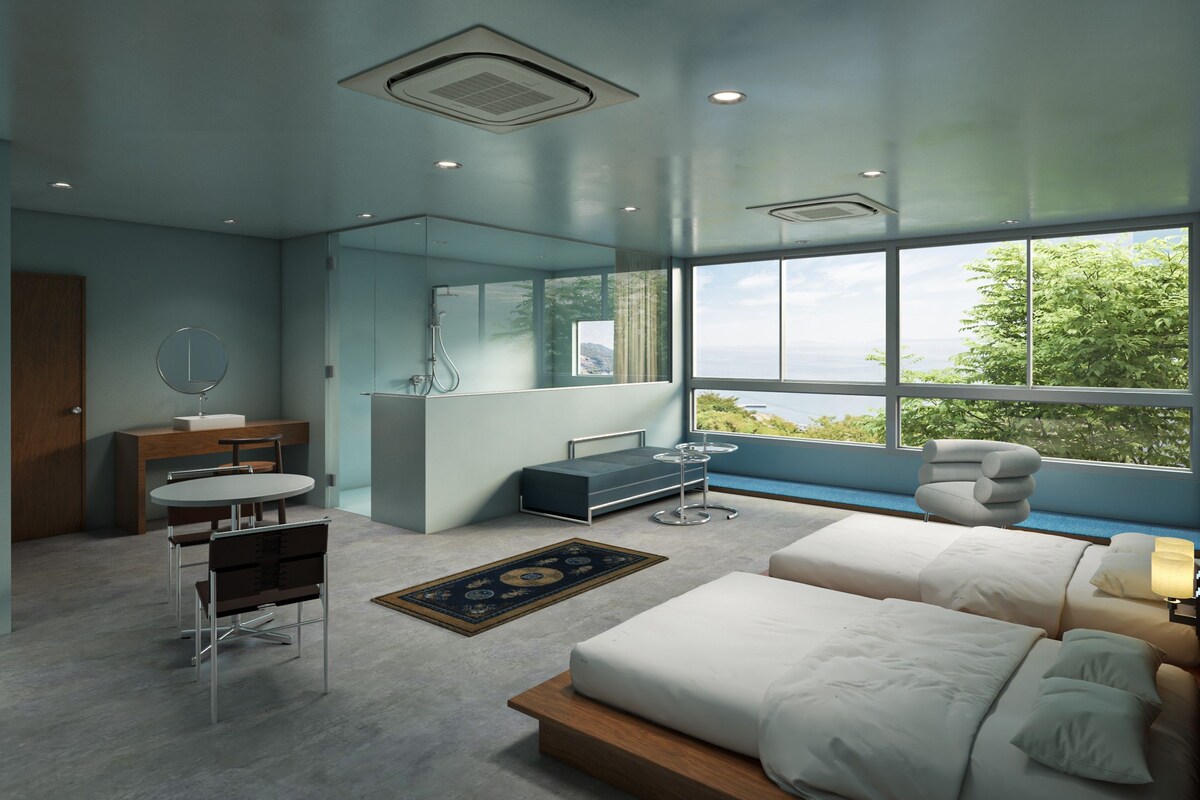 A modern bedroom with a large bed, a sitting area with a table and chairs, a vanity with a mirror, and a view of the outdoors through large windows.