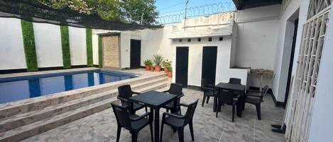 Un área de piscina con suelo de baldosas, mobiliario exterior y una sección cubierta con techo.