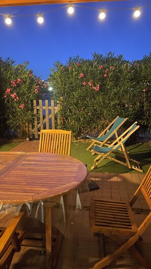 Een terras buiten met een houten tafel en stoelen, omringd door een houten hek en bloeiende struiken.
