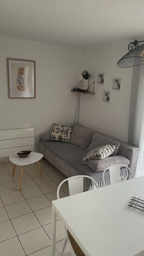 Ett minimalistiskt vardagsrum med en grå soffa, ett litet runt bord, en vit stol och en vägghylla med en lampa.