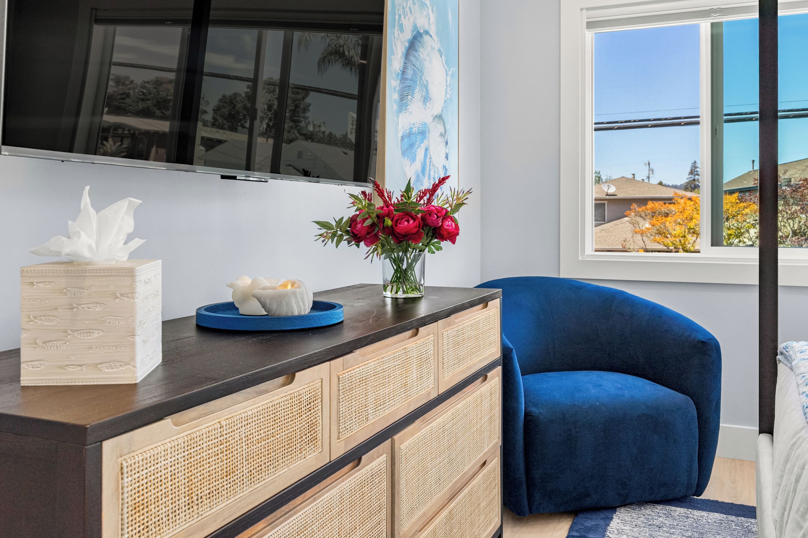 Una sala de estar moderna con un sofá azul oscuro, un mueble de madera con puertas tejidas, un televisor de pantalla plana montado en la pared y una ventana con vistas a casas y árboles.