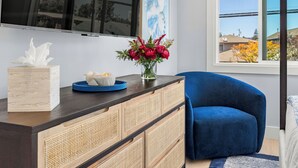 Een moderne woonkamer met een donkerblauwe bank, een houten kast met gevlochten deuren, een flatscreen televisie aan de muur en een raam met uitzicht op huizen en bomen.