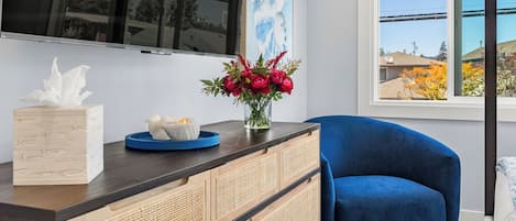 Una sala de estar moderna con un sofá azul oscuro, un mueble de madera con puertas tejidas, un televisor de pantalla plana montado en la pared y una ventana con vistas a casas y árboles.