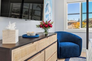A modern living room with a dark blue sofa, a wooden cabinet with woven doors, a flat-screen TV mounted on the wall, and a window with a view of houses and trees. - Santa Cruz Escape-Rooftop Deck/Ocean Views/Hot Tub (Santa Cruz)