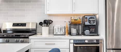 Une cuisine moderne dotée d’une cuisinière, d’un four et d’un réfrigérateur en acier inoxydable, de meubles blancs et d’une machine à café.