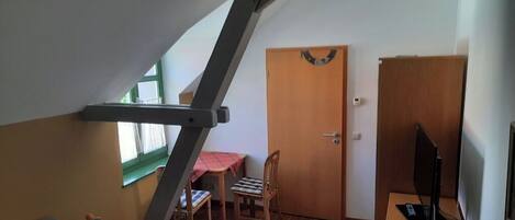 A compact room with a sloped ceiling, a desk with a computer, a chair, a small table with a lamp, and a door.