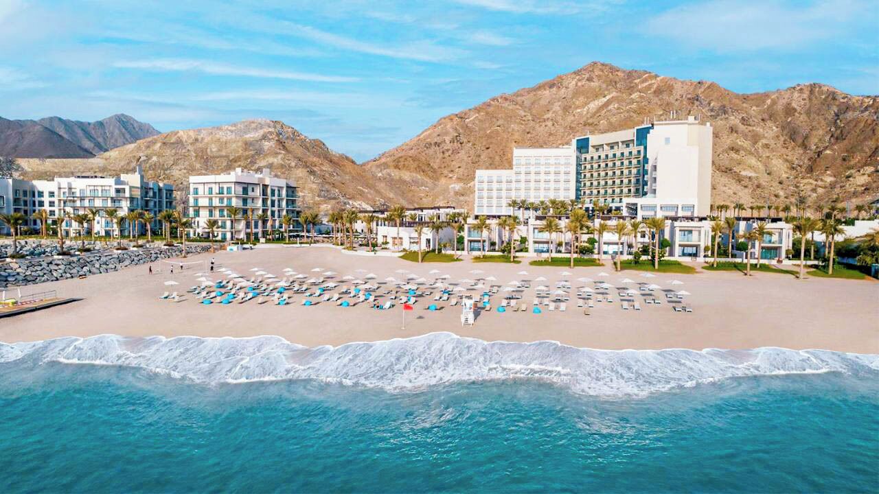 A beachfront hotel with multiple buildings, a sandy beach, and a mountain backdrop.
