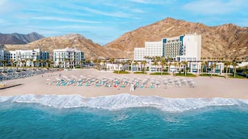 A beachfront hotel with multiple buildings, a sandy beach, and a mountain backdrop.