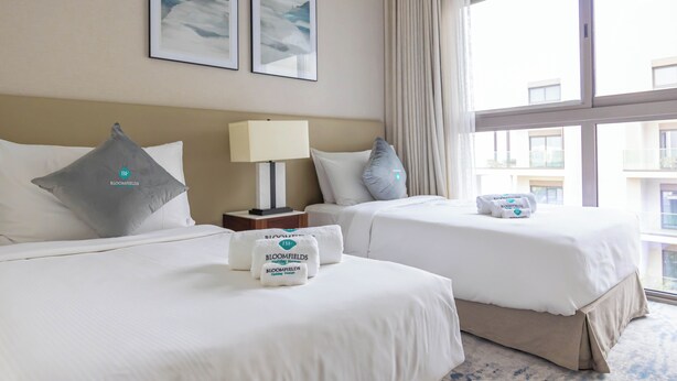 A hotel room with two beds, a nightstand, a lamp, and a window with a view of buildings. - Bloomfields 2BR with maid Address Resid (Al Aqah)