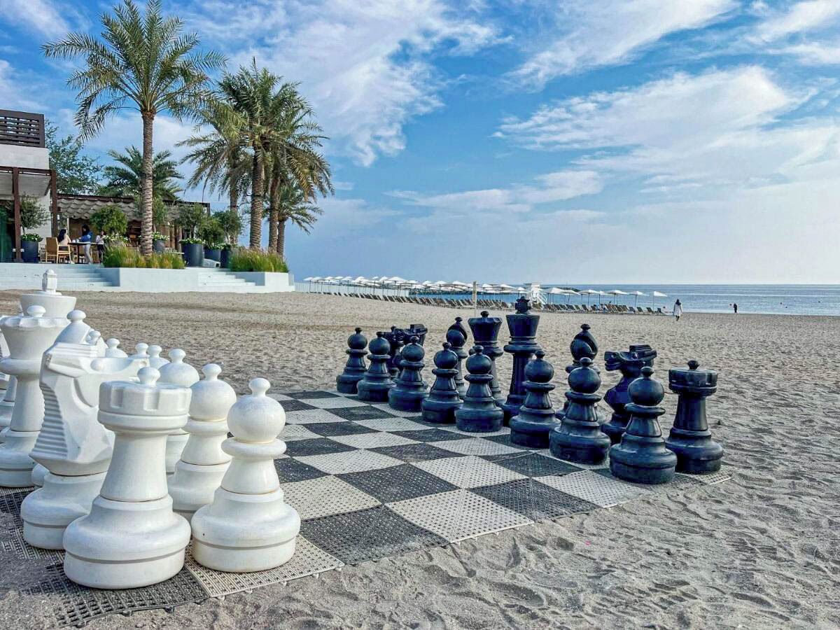 A chessboard set up on a sandy beach with palm trees and a building in the background.
