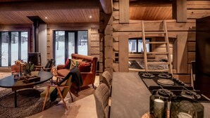 En hyggelig stue med pejs, et spiseområde med komfur og store vinduer med udsigt til naturen.