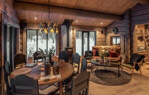 Ein gemütliches Blockhauss-Interieur mit Essbereich, einer Sitzecke mit Kamin und großen Fenstern mit Blick auf eine verschneite Landschaft.